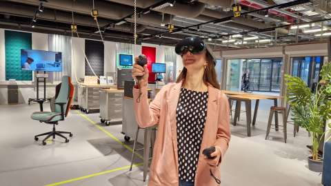Eine Frau trägt eine Virtual Reality-Brille und benutzt zwei Controller, während sie sich dabei durch einen Raum bewegt.