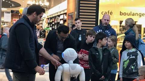 Das Bild zeigt Erwachsene und Kinder vor einem Geschäft wie sie mit dem Roboter interagieren.