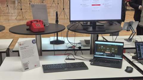 Physisiche Präsenz des Demonstrator KI im Büro. Aus dem Bild sind ein Bildschirm, Tastatur, Laptop, Maus und ein rotes Telefon.