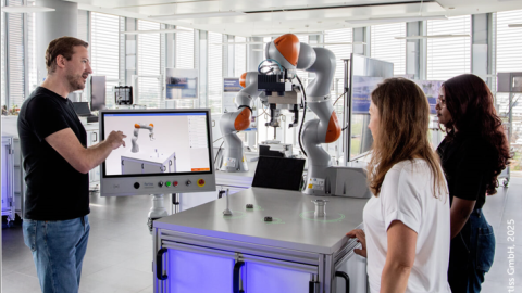 Der Cobot steht auf einem Tisch in der Bildmitte. Links präsentiert ein Mitarbeiter seine Arbeitsweise. Zwischen dem Cobot und dem Mitarbeiter zeigt ein Bildschirm eine Simulation des Cobots. Rechts hören zwei Besucher den Erläuterungen zu.