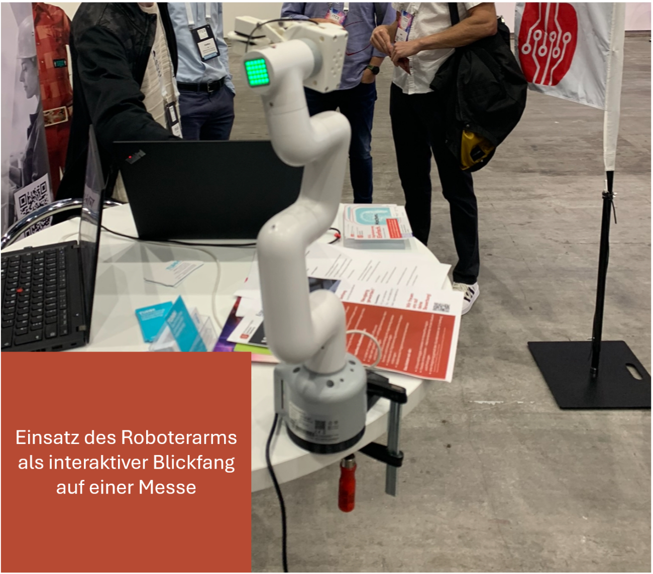 Ein weißer Roboterarm ist an einem Messestand auf einem Tisch befestigt. Der Roboterarm dient als interaktiver Blickfang und demonstriert automatisierte Bewegungsabläufe.