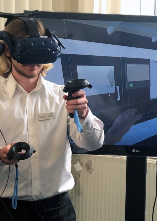Nutzer mit VR-Headset und Controllern vor einem Bildschirm, auf dem die VR-Umgebung zu sehen ist.