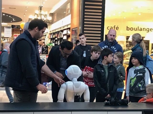 Das Bild zeigt Erwachsene und Kinder vor einem Geschäft wie sie mit dem Roboter interagieren.