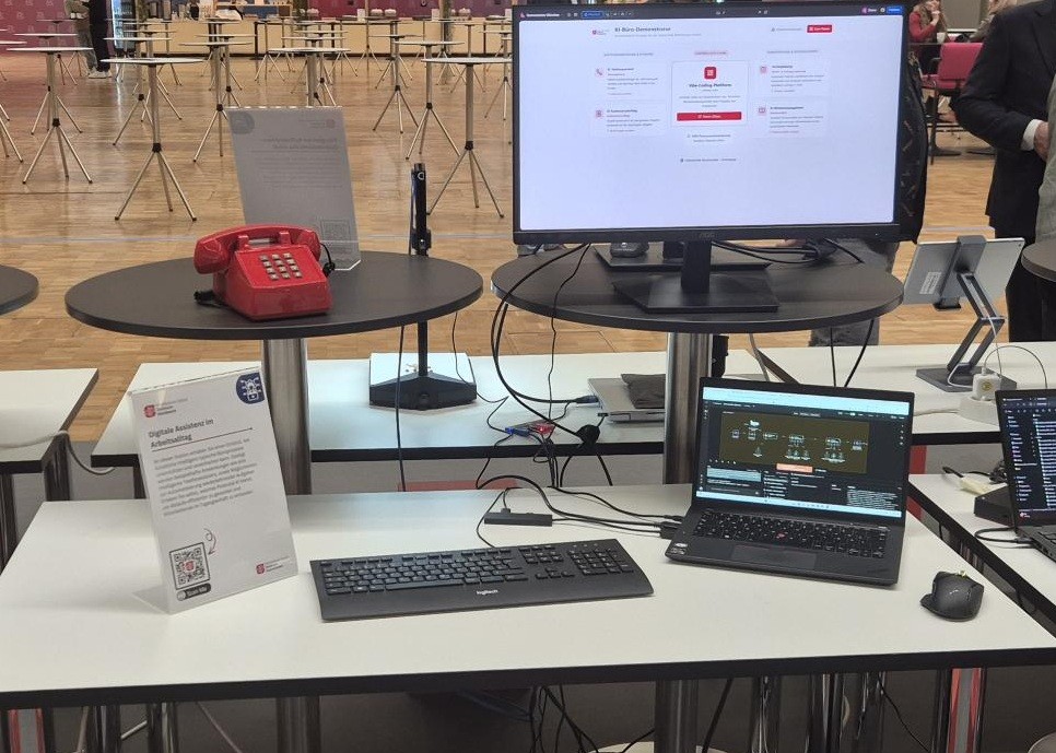 Physisiche Präsenz des Demonstrator KI im Büro. Aus dem Bild sind ein Bildschirm, Tastatur, Laptop, Maus und ein rotes Telefon.